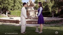 a man and a woman are standing in front of a fountain with the good place written on the bottom