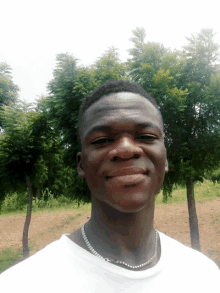 a young man wearing a white shirt and a necklace smiles in front of some trees