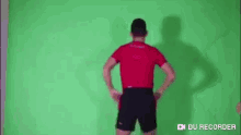 a man in a red shirt and black shorts is dancing in front of a green screen .