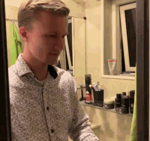 a man in a white shirt stands in front of a mirror in a bathroom with a shelf full of beauty products