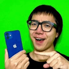 a man wearing glasses is holding a blue iphone and pointing at it on a green screen .