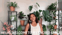 a woman stands in front of a display of potted plants and says " hello welcome to my sacred space "