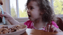 a little girl is sitting at a table with a plate of food and a fork in her hand .