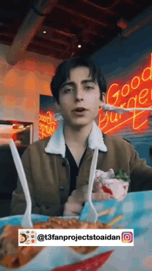 a young man is sitting at a table with a plate of food in front of a neon sign that says good burger .