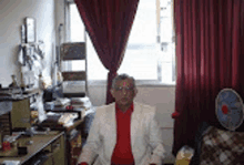 a man in a white jacket and red shirt is sitting in a room with red curtains