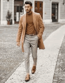 a man is walking down a sidewalk wearing a tan coat , brown turtleneck , and grey pants .