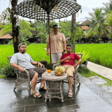 three men are sitting under an umbrella in front of a field of rice