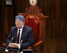 a man in a suit and tie is sitting in a wooden chair in front of microphones .