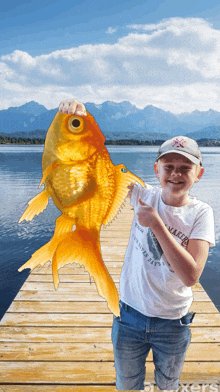 a boy holding a large goldfish on a dock wearing a shirt that says maker