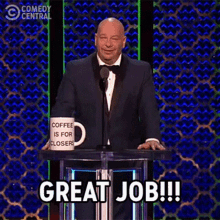 a man in a tuxedo is giving a speech at a podium with a coffee mug in front of him .