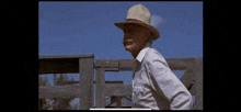 an older man wearing a hat and a white shirt is standing in front of a wooden fence .