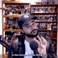 a man wearing a ny hat is talking into a microphone in front of a shelf full of toys .