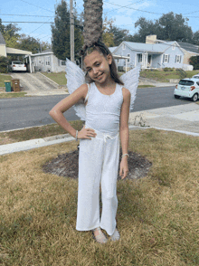 a young girl in a white angel costume stands in the grass with her hands on her hips