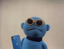 a blue puppet wearing sunglasses is holding an apple iphone .