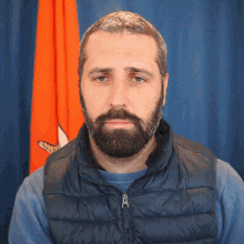 a man with a beard wearing a blue shirt and a black vest stands in front of an orange flag