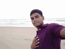 a young man in a purple shirt stands on a beach with his hand on his chest