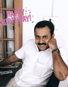 a man in a white shirt sits in front of a sign that says " happy birthday "