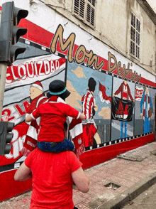 a man is carrying a child on his shoulders in front of a mural that says mon destin