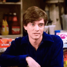 a young man in a blue shirt is sitting at a table in front of a shelf with cereal on it .