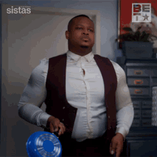 a man in a white shirt and a maroon vest is holding a blue bowl in front of a sign that says sistas