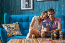 a man and woman are sitting on a blue couch holding a box that says ' happy birthday '