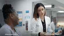 a woman in a lab coat is talking to another woman in a hospital hallway with the nbc logo in the corner