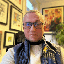 a man wearing glasses and a blue vest is standing in front of a wall of framed pictures