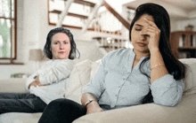 a woman sits on a couch with her hand on her forehead while another woman sits behind her