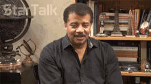 a man in a black shirt is sitting in front of a bookshelf with the word talk written on the wall behind him