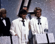 a group of men in white jackets and bow ties stand in front of a microphone