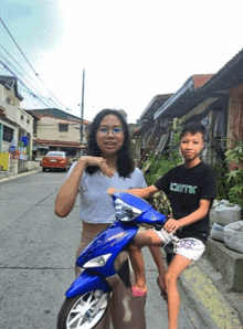 a girl is sitting on a blue scooter next to a boy wearing a shirt that says ibetk