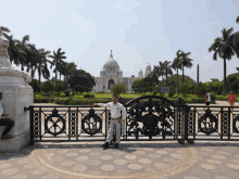 a man stands in front of a gate with a large white building in the background