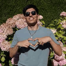 a man wearing sunglasses and a blue shirt is making a heart shape with his hands in front of pink flowers .