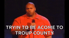 a man in a red shirt stands in front of a microphone with the words tryin to be a come to troup county below him