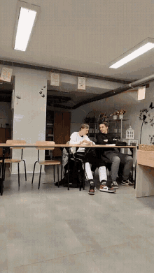 two boys sit at a table in a classroom with a sign on the wall that says ' i love you '