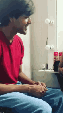 a man in a red shirt sits in front of a mirror with a bottle of salt on the counter