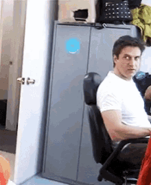 a man in a white shirt is sitting in an office chair in front of a cabinet with a blue sticker on it