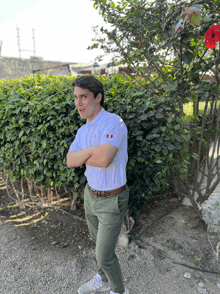 a man in a white polo shirt stands with his arms crossed in front of a bush