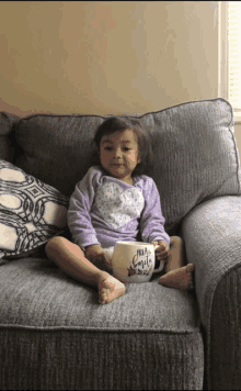 a little girl is sitting on a couch holding a mug that says hola emele