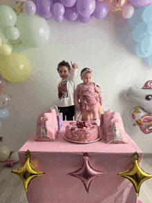 two children standing in front of a pink table with balloons and a cake on it