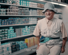 a man wearing a hat and vest is sitting in front of a grocery aisle with hebrew writing on the bottom