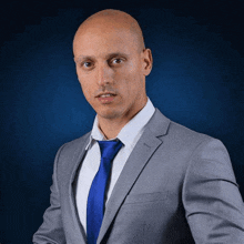 a bald man is wearing a suit and tie and standing in front of a blue background .