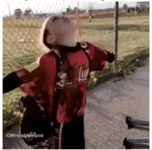 a young boy in a red shirt is standing in front of a chain link fence with his arms outstretched .