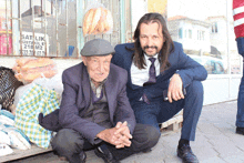 a man in a suit sits next to an older man in front of a sign that says " satilik "