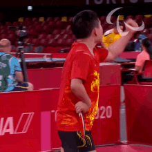 a young boy in a red shirt is holding a racket in front of a wall that says tokyo 2020