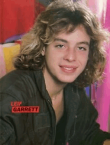 a young man with long curly hair is smiling and wearing a black jacket .