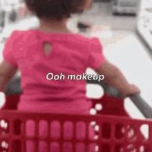 a little girl is sitting in a shopping cart with the words `` ooh makeup '' written on her back .