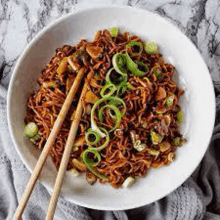 a white bowl filled with noodles , mushrooms , and green onions with chopsticks .