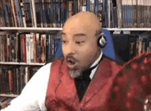 a bald man wearing headphones and a red vest is sitting in front of a bookshelf in a library .