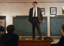 a man in a suit and tie stands on a desk in a classroom